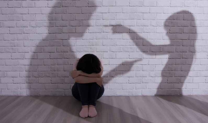 Une femme recroquevillée avec la tête dans ses bras. Des ombres sur le mur se pointent mutuellement du doigt
